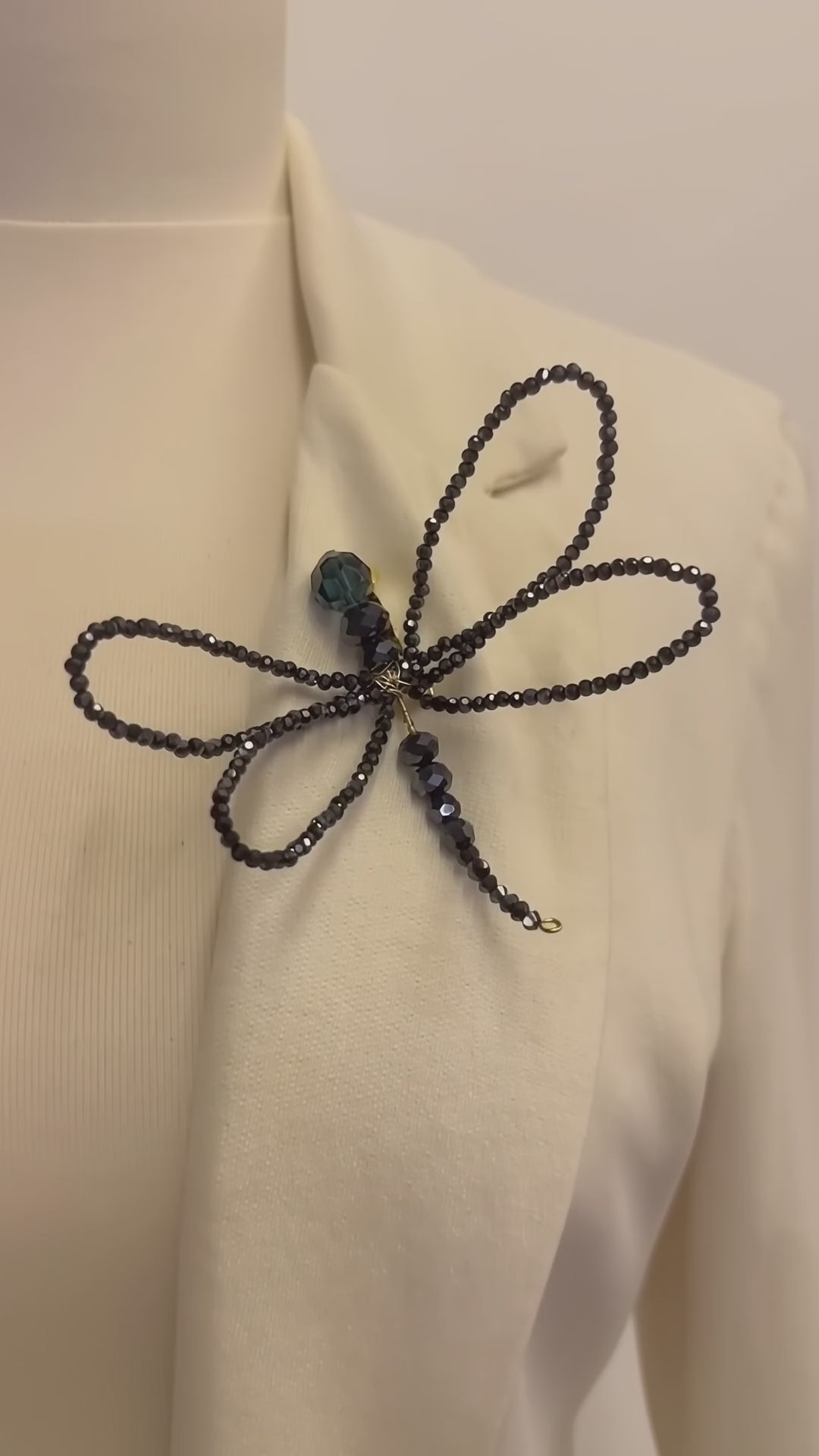 Handmade LUMI black crystal dragonfly brooch with four delicate beaded wings and a long beaded tail, displayed on an ivory blazer in a clean white studio.